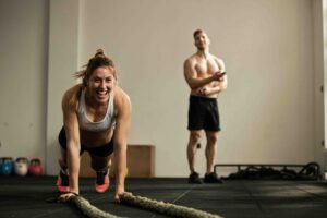 Cheerful female athlete doing push-ups while exercising with battle rope and having fun in a gym There is a man in the background.